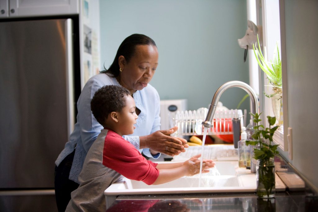A woman teaches her son how to wash his hands at their kitchen sink